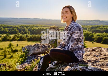 Smiling young woman hiker sitting on rock in mountains enjoying prefect summer weather Stock Photo