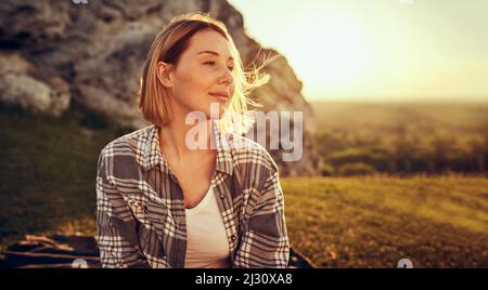 Thoughtful young woman sitting on meadow during hike on sunny evening Stock Photo