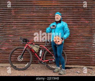 senior male cyclist with a gravel bike is taking a break at old corrugated metal barn in Colorado coutryside Stock Photo