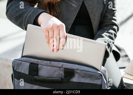 Crop anonymous female freelancer in outerwear sitting on concrete bench and taking netbook from case for working remotely Stock Photo