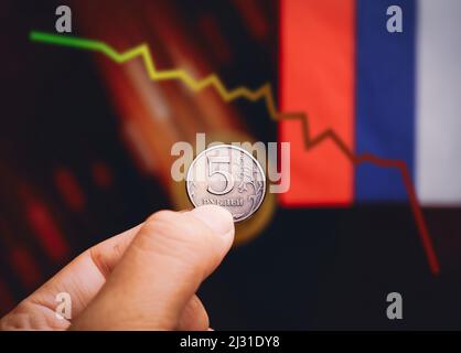 Russian ruble coin holding on the hand with falling down trading charts and russian flags blurred background Stock Photo