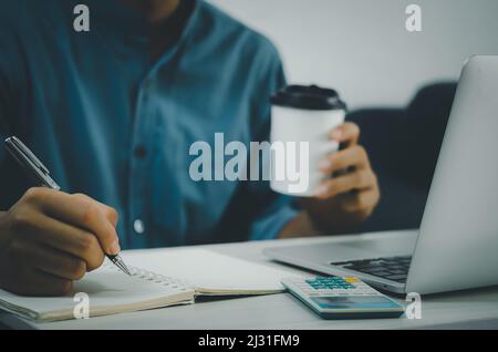 Male hand holding pen and writing at notebook and coffee cup on desk with notebook computer and calculator. Stock Photo