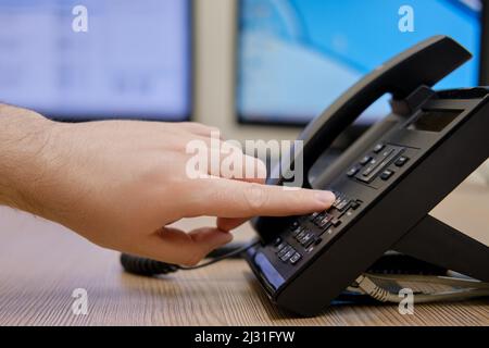 A man hand dials a number on the phone at the monitors of office computers. Finger presses on the buttons of a office landline phone Stock Photo