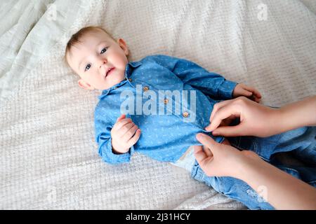 A mother woman wears a blue shirt to a baby boy. Mom dresses a happy child in clothes on the bed. Stock Photo