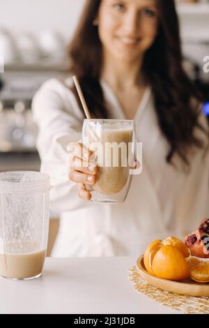 A happy smiling healthy woman has prepared a fruit cocktail and gives it to you while standing at home in the kitchen.Healthy eating. Stock Photo