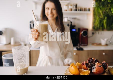 A happy smiling healthy woman has prepared a fruit cocktail and gives it to you while standing at home in the kitchen.Healthy eating. Stock Photo