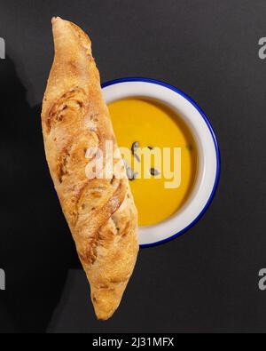 Home made pumpkin soup puree with seeds in a blue plate with fresh crunchy french baguette on a black background. Vegan vegetable soup puree with loaf Stock Photo