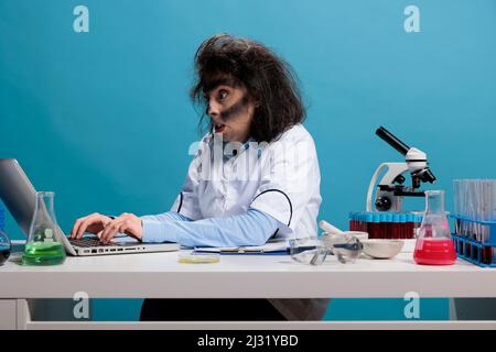 Insane angry biochemist with lunatic look after lab explosion sitting at desk and reviewing chemical experiment report. Wild foolish woman with smokey face researching discovery on laptop. Studio shot Stock Photo