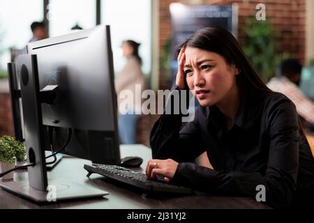 Doubtful asian computer engineer puzzled by nonfunctional security algorithm and company corrupted database. Confused uncertain programmer sitting at computer trying to solve network processing errors Stock Photo