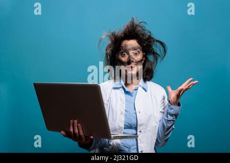 Lunatic chemist with messy hair and dirty face having laptop while gesticulating angrily after dangerous lab explosion. Insane scientist being enraged after failed laboratory chemical experiment. Stock Photo