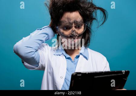 Wacky and silly looking crazy chemist with touchscreen tablet, puzzled by failed experiment report. Mad scientist with messy hair and dirty face looking confused after dangerous chemical explosion. Stock Photo