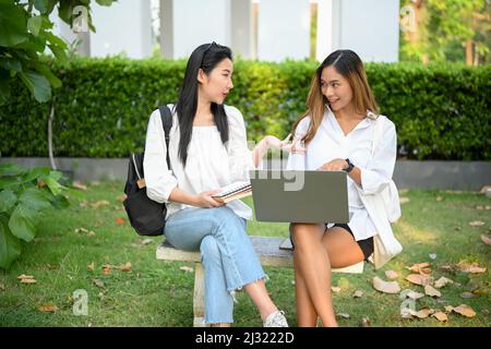 Two Asian young female college students sitting in the park, discussing their project and searching some informations on the internet via laptop compu Stock Photo