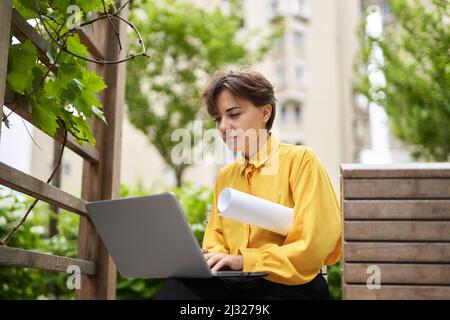 Successful smiling business lady in yellow blouse sitting on bench outdoor in park with plans using laptop working or having online meeting. Business, finance industry or real estate concept Stock Photo