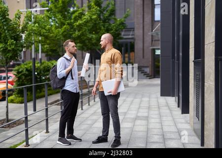 Male real estate agents talking outdoor with blueprints and laptop. Construction, architecture or design managers concept. On the go meeting in downtown. High quality image Stock Photo