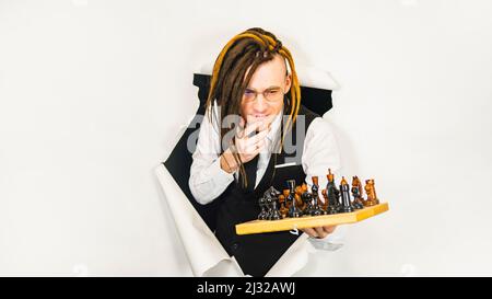 Young man in glasses with dreadlocks plays chess in hole of white background. Handsome male in white shirt, black vest and tie playing in logical boar Stock Photo