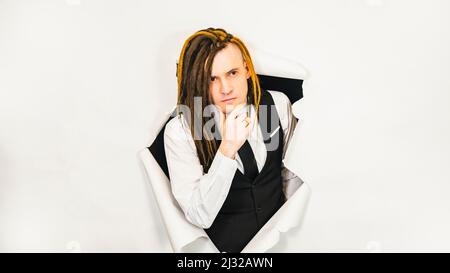 Young man with dreadlocks scratching chin. Handsome male in white shirt, black vest and tie with thoughtful look in hole of white background Stock Photo