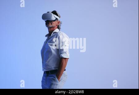 Senior businessman wearing a virtual reality headset in a studio. Mature businessman looking away while standing with his hands in his pockets. Busine Stock Photo