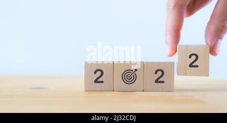 Business goals in 2022 concept. Businessman hand holds wooden cubes '2022' with 'dart board has dart arrow hitting' icon on beautiful  background and Stock Photo