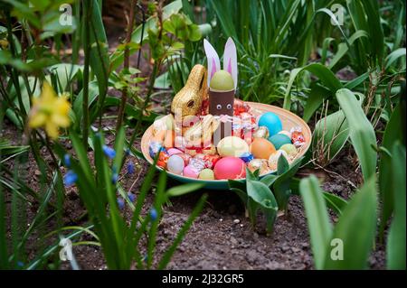 PRODUCTION - 01 April 2022, Berlin: ILLUSTRATION - An Easter nest with made bunny, chocolate bunny, colored eggs and chocolate eggs lie in a flower bed. (posed scene) Photo: Annette Riedl/dpa Stock Photo
