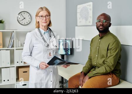 Confident experienced female doctor in whitecoat and African American patient in casualwear looking at camera in medical office Stock Photo