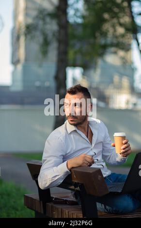Thoughtful man in the park with a laptop drinking coffee. A young man on a background of green trees, a hot sunny summer day. Warm soft light, close-up. Stock Photo