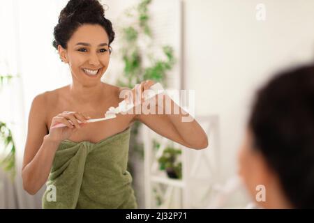 Attractive young woman with lovely smile wearing towel after shower, applying toothpaste on brush near mirror at home Stock Photo