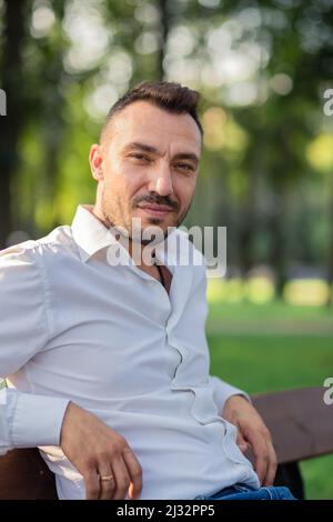 A happy business man in a white shirt is resting while sitting on a bench. Young man in city park, warm sunny summer day. Warm soft light, close-up. Stock Photo