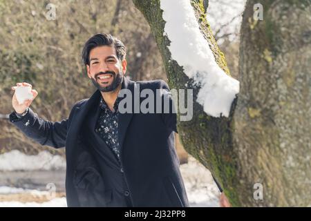 Cheerful Stylish Hispanic Latin Man dressed in a black camel coat throwing the snowball to the camera - winter leisure concept. High quality photo Stock Photo
