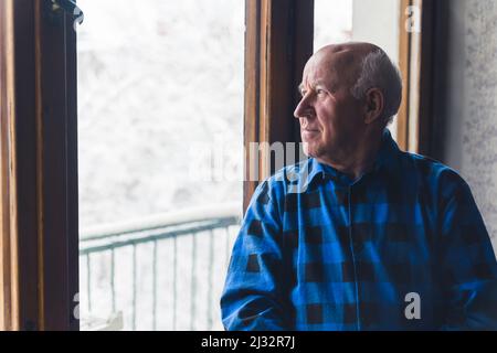 Sad Caucasian Bald Senior Pensioner wearing blue checked shirt Looking through the window with winter scenery - Sadness concept. High quality photo Stock Photo