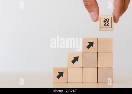 Business growth in 2022 concept. Businessman holds wooden cubes with text '2022' standing on increasing arrow icon on  white background. Goals, vision Stock Photo