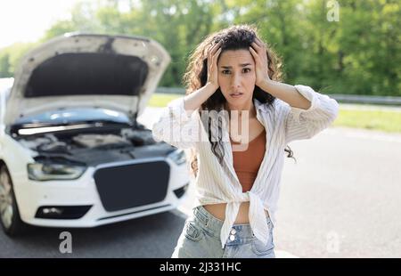 Young stressed woman standing on highway near broken car, grabbing head in terror, waiting for breakdown road service Stock Photo