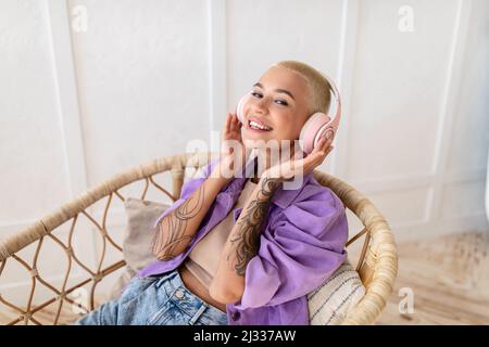 Young lady in wireless headphones chilling in wicker armchair, listening to music and smiling at camera at home Stock Photo