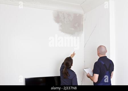 Owner showing a repairman or plumber a big water stain and various cracks in wall and ceiling of her living room in an old building. Stock Photo