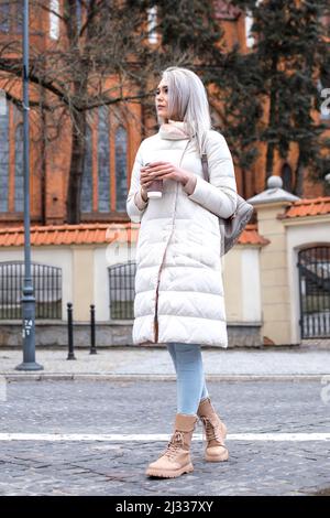 Portrait of young attractive pensive woman with long grey hair holding takeaway cup of coffee, standing in white jacket. Stock Photo