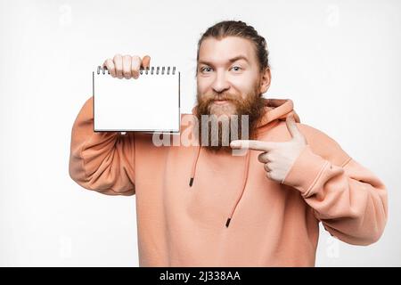 Scandinavian handsome happy man holding notepad and points to paper mockup isolated on gray background. Guy with ginger hairstyle and beard in a sweat Stock Photo