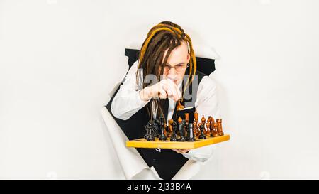 Young man in glasses with dreadlocks plays chess in hole of white background. Handsome male in white shirt, black vest and tie playing in logical boar Stock Photo