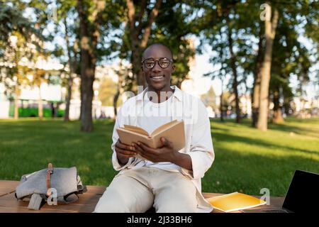 Happy black student guy reading interesting book, having rest after classes in park, sitting on bench on sunny day Stock Photo
