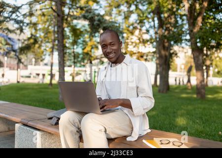 Rest at campus. Happy african american male college student relaxing outdoors with laptop, sitting on bench in park Stock Photo