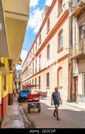 A Cuban man wearing a face mask walks in a narrow street in Old Havana. There is an empty bicitaxi or pedicab parked. Traditional architecture of buil Stock Photo