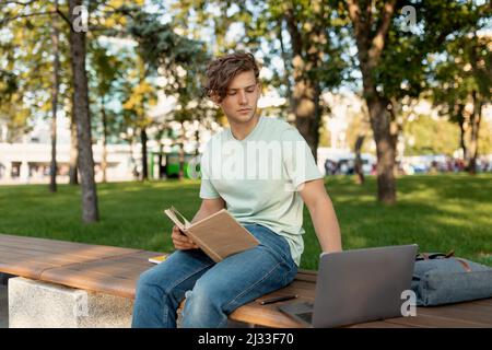 Focused caucasian student guy doing homework with laptop and book, sitting on bench in park or college campus Stock Photo
