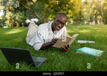 Happy black student guy in eyeglasses reading book, preparing for exams and enjoying studying outdoors in park Stock Photo