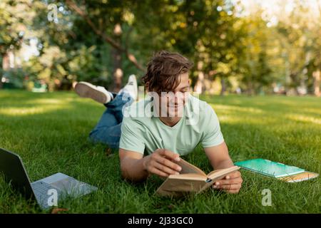 Cheerful caucasian student guy reading book outdoors, preparing for lectures while lying on lawn in university campus Stock Photo
