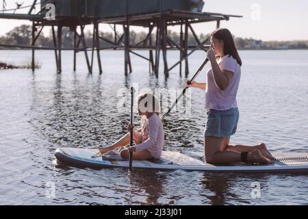 Little girl with mother sup boarding together on one sup in daytime with house on water in background. Active lifestyle. Time together. Teaching Stock Photo