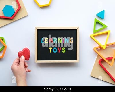 Text Learning Toys on blackboard. Nested wood triangles and hexagons in rainbow colors. Red wooden heart in human hand. Stock Photo