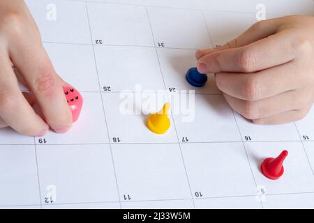 Board game and children's leisure concept - a child's hand moves a game chip on the field and holds a cube with numbers. Multi-colored chips on a line Stock Photo