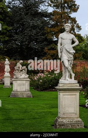 Neoclassical statues in the Rose Garden at Hampton Court, historic royal palace beside the River Thames in the London Borough of Richmond-upon-Thames, England:  Adonis, Greek god of beauty and desire, hand casually resting on hip; a loving mother preparing to breastfeed her baby in ‘Abundance’ by British sculptor, Francis Derwent Wood (1871 - 1926); and (background) Flora, Roman goddess of flowers and spring. Stock Photo