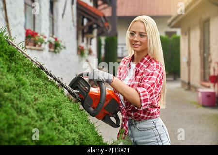 Beautiful caucasian woman in casual wear, safety glasses and gloves using petrol hedge trimmer for cutting bushes. Competent female gardener working outdoors.  Stock Photo