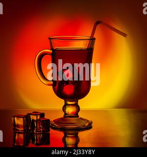 Cold tea, cocktail, fruit drink in a tall glass cup with a drinking straw on a table with pieces of ice on a beautiful bright yellow-red gradient back Stock Photo