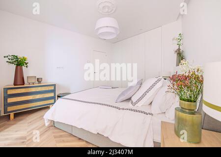 Bedroom with double bed with cushions in light tones, light wood bedside tables, wicker chest of drawers and vases with decorative flowers Stock Photo