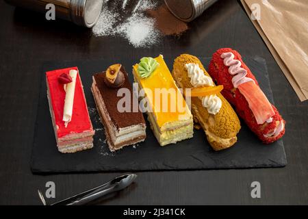 Assorted cakes and pastries with chocolate and fruit glazed with creams on black slate tray Stock Photo